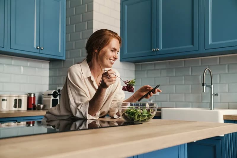 Mulher comendo distraída ao celular, ilustrando conteúdo sobre engordar depois dos 40.