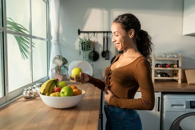 Mulher escolhendo uma maçã em uma cozinha iluminada, ilustrando hábitos alimentares na manutenção do peso.