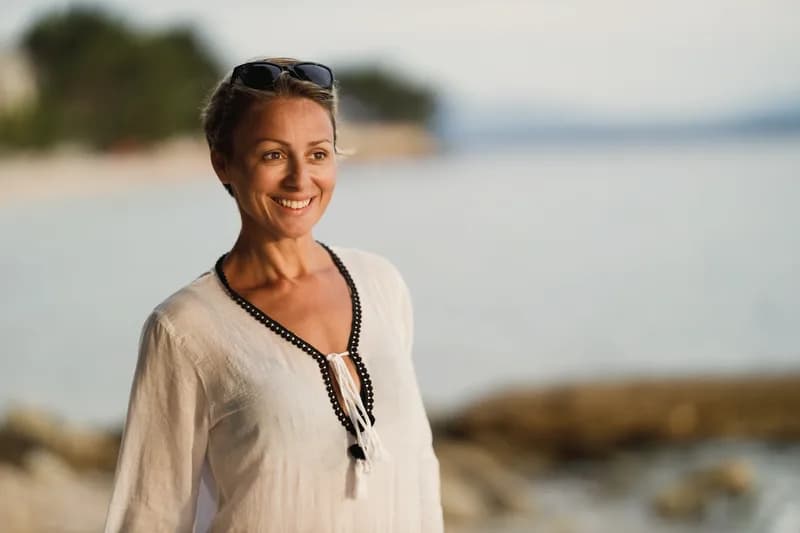 Mulher de 40 e poucos anos, serena e confiante, sorrindo à beira-mar ao entardecer.